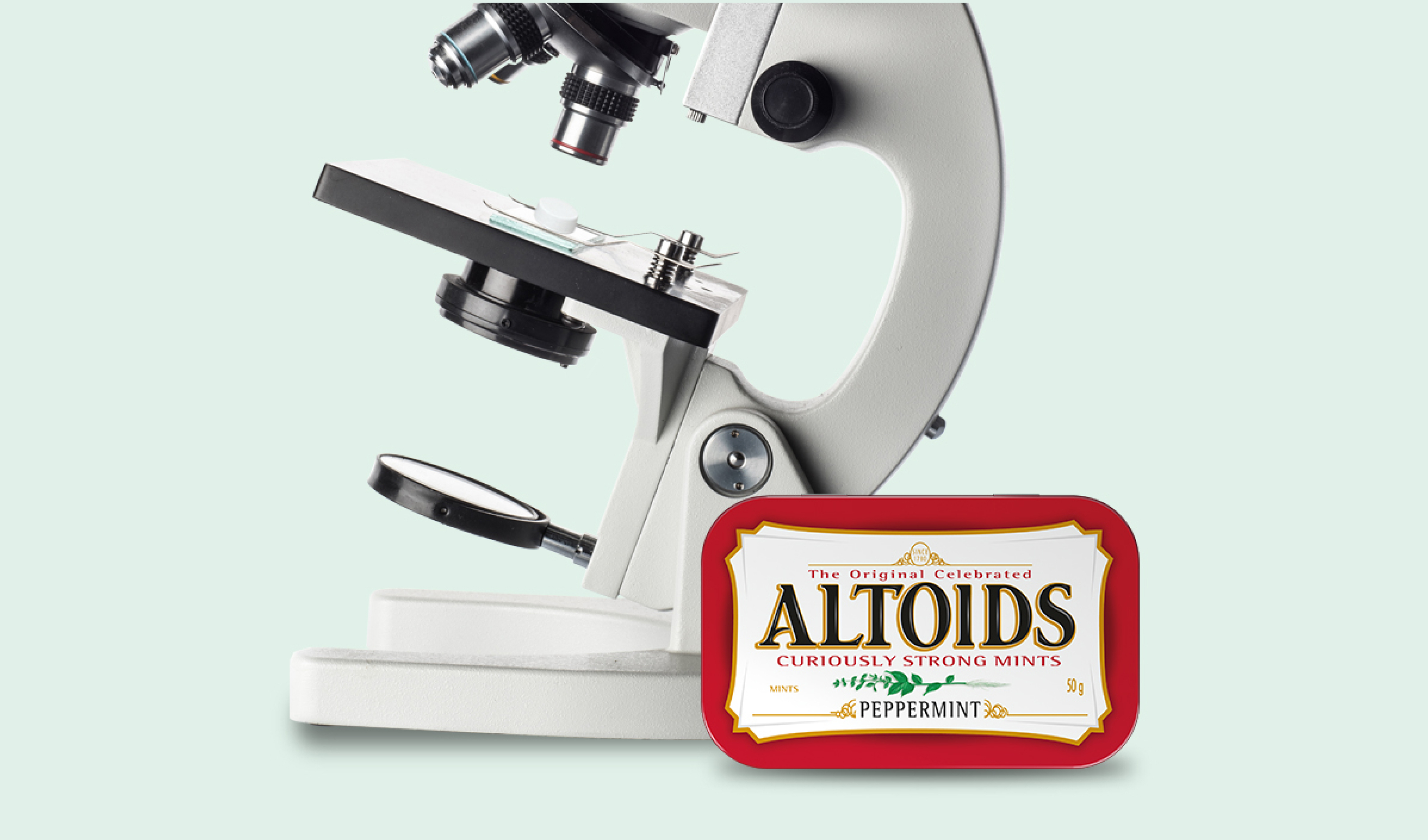Tin of Peppermint Altoids in front of a microscope on a mint green background