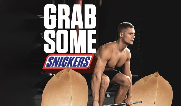 Snickers advertisement showing shirtless man picking up weights made of giant peanuts
